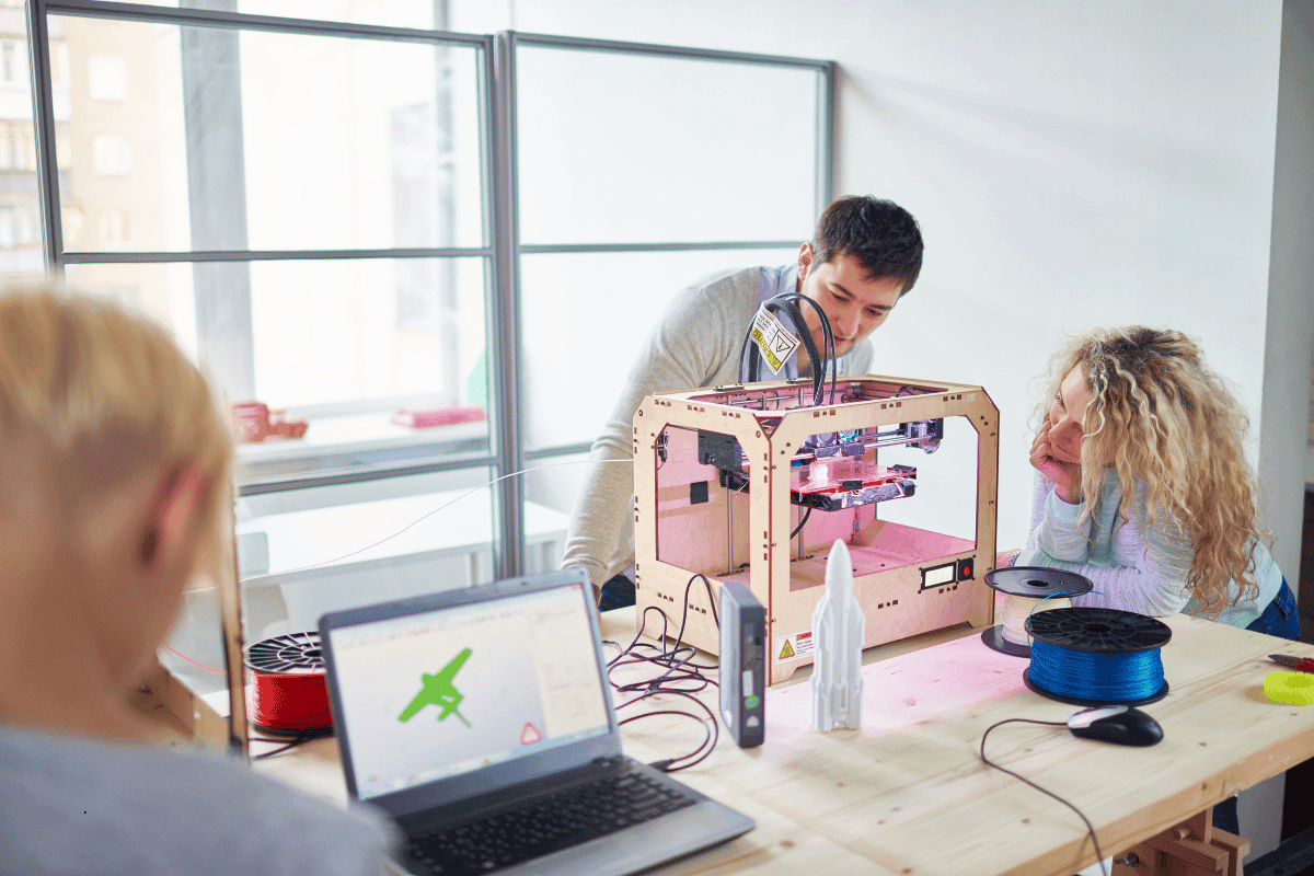 Engineering students using a 3D printer and CAD software to develop a white rocket prototype.
