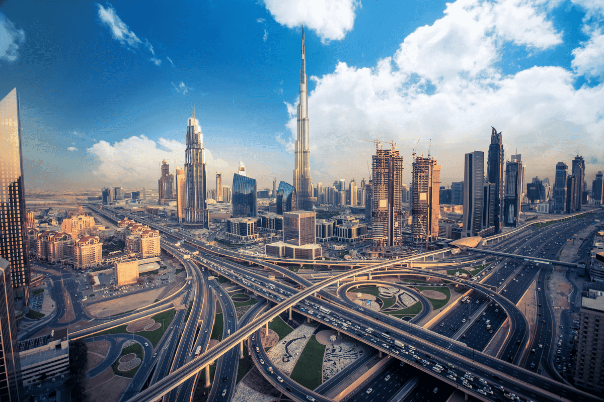 Aerial view of the Burj Khalifa and Dubai skyline with complex highway interchanges and corporate skyscrapers under a blue sky.